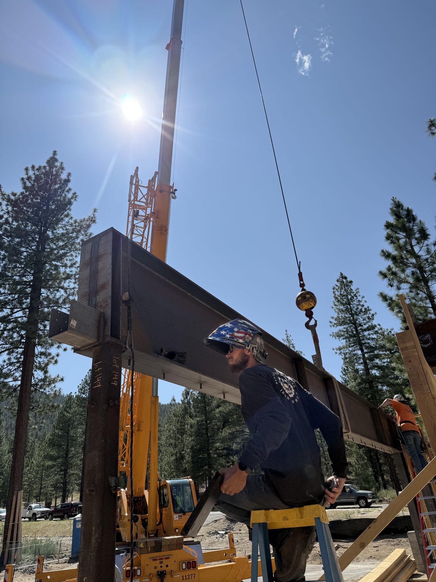 Crane-assisted steel beam installation with worker on ladder at custom home site